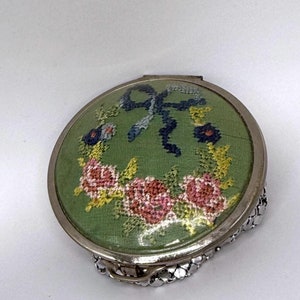 以下が含まれることがあります： 緑色の布地にピンクのバラと青いリボンが刺繍された、丸い銀色のコンパクト