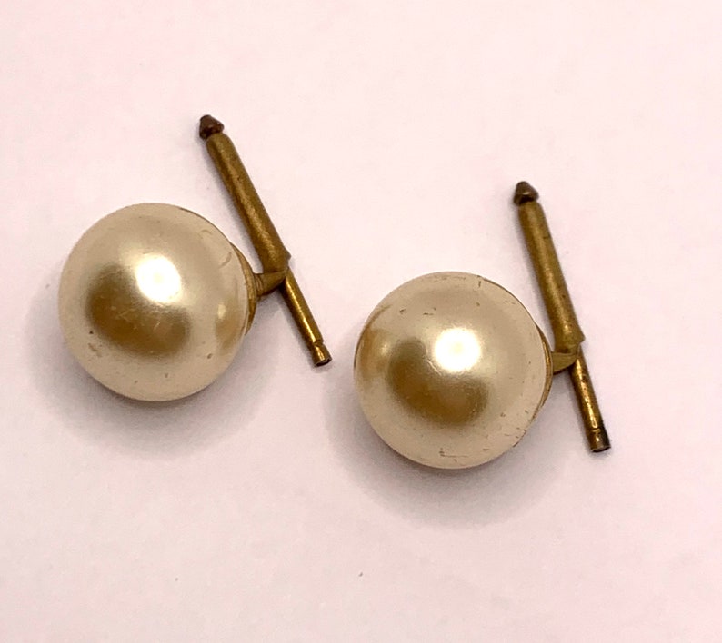 May include: A pair of gold-tone earrings with faux pearl accents. The pearls are round and off-white in colour.