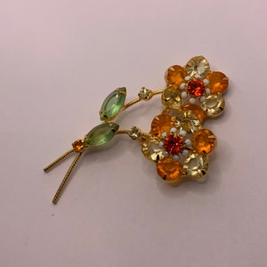May include: A gold-tone metal brooch featuring two flower blossoms with orange and pale yellow faceted crystals, white enamel accents, and two light green marquise-cut crystal leaves on textured stems with small clear crystal embellishments.