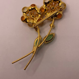 May include: A gold-tone metal flower brooch featuring two blossoms with amber and clear faceted stones. The stems are textured and include two light green marquise-cut leaves. A horizontal pin mechanism is attached to the back of the flower heads.