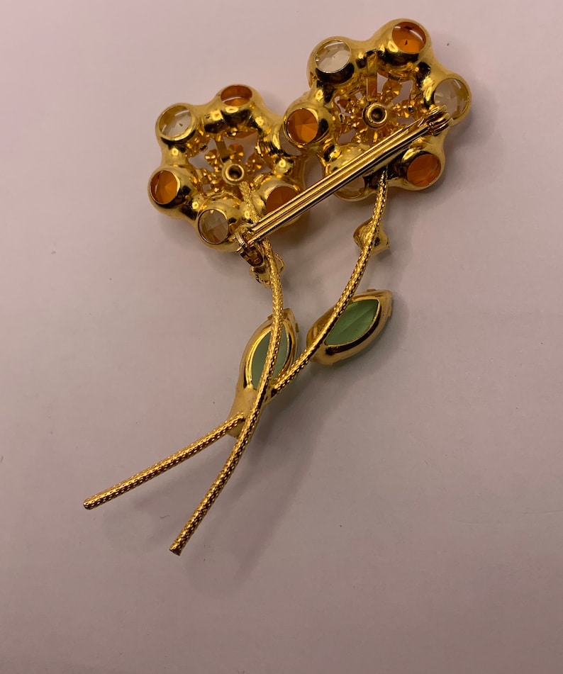 May include: A gold-tone metal flower brooch featuring two blossoms with amber and clear faceted stones. The stems are textured and include two light green marquise-cut leaves. A horizontal pin mechanism is attached to the back of the flower heads.