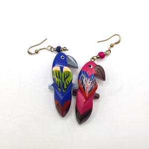 Op de afbeelding: Handgeschilderde houten papegaai oorbellen. De ene oorbel is blauw met groene en gele accenten, de andere is roze met rode en blauwe details. Beide hebben grijze snavels en goudkleurige oorhaakjes met kralen.