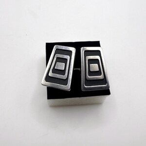 May include: A pair of silver-toned earrings with a geometric design. Each earring features a rectangular shape with three nested squares in a black background. The earrings are displayed on a black surface against a white background.