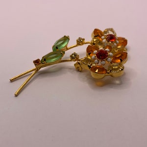 May include: A gold-tone metal brooch shaped like two flowers with orange and yellow faceted crystals, white enamel accents, and two light green marquise-cut crystal leaves on thin textured stems with small clear crystal embellishments.