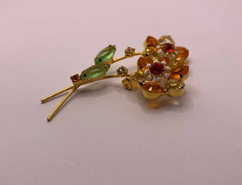 May include: A gold-tone metal brooch shaped like two flowers with orange and yellow faceted crystals, white enamel accents, and two light green marquise-cut crystal leaves on thin textured stems with small clear crystal embellishments.