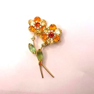 May include: A gold-tone metal brooch shaped like two flowers with stems. The flower petals are made of faceted orange, yellow, and clear crystals with a central red crystal. Two light green marquise-shaped crystals form the leaves on the stems.