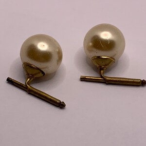 May include: A pair of cufflinks with a pearl-like white sphere on top of a gold-coloured base. The base has a swiveling arm that connects to a gold-coloured bar.