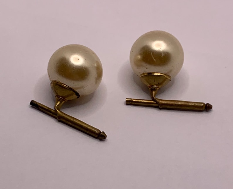 May include: A pair of cufflinks with a pearl-like white sphere on top of a gold-coloured base. The base has a swiveling arm that connects to a gold-coloured bar.