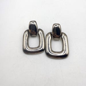 May include: A pair of silver-toned, rectangular drop earrings. Each earring features a rounded top and a large, open rectangle below. The earrings are displayed on a white background, showcasing their polished, metallic finish. The design is simple and modern.