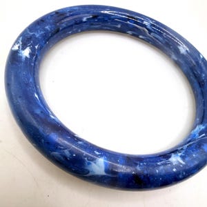 Puede incluir: Un brazalete circular con acabado pulido y un patrón jaspeado en tonos azul profundo, azul marino, blanco y pequeños acentos negros a lo largo de la superficie lisa y redondeada de la pieza de joyería.