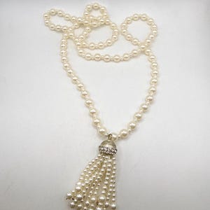 Puede incluir: Un collar de perlas de color crema con un colgante de borla. El collar presenta una hebra de pequeñas perlas redondas. El colgante tiene una tapa plateada adornada con pequeños cristales transparentes y una borla de perlas.