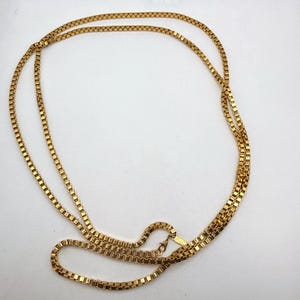 Könnte beinhalten: Eine goldfarbene Halskette mit einem Box-Chain-Design. Die Halskette liegt auf einer weißen Oberfläche. Die Kette besteht aus kleinen, quadratischen Gliedern, die ein strukturiertes Aussehen erzeugen. Die Halskette hat einen Verschluss.