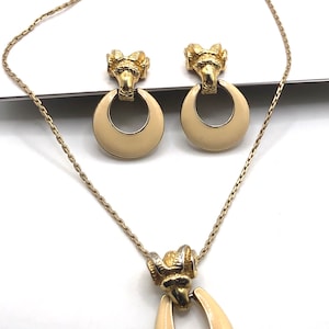 Puede incluir: Un conjunto de collar y pendientes en tono dorado con un colgante y pendientes de esmalte crema. El colgante y los pendientes presentan un diseño en tono dorado con un anillo en la parte inferior.