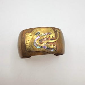 Op de afbeelding: Een brede, koperkleurige manchetarmband met een goudkleurig, gestileerd drakenontwerp. De kenmerken van de draak zijn ingelegd met iriserende abalonschelp. De armband heeft een rechthoekige vorm met afgeronde randen.