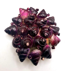 May include: A circular brooch composed of numerous small, spiraled sea shells dyed in deep shades of purple and magenta. The shells are clustered tightly together, creating a textured, dome-like surface with a dark, glossy finish throughout.