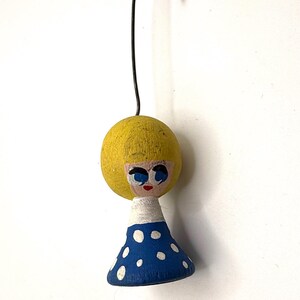 May include: A vintage-style wooden doll ornament with a yellow head, blue eyes, and a red mouth. The doll has a blue dress with white polka dots and a white collar. A black wire loop is attached to the top for hanging.