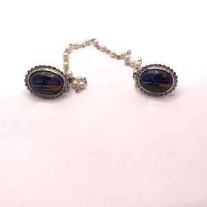 Op de afbeelding: Een paar zilverkleurige ovale broches met een blauw en oranje vlinderontwerp. De broches zijn verbonden door een ketting van nepparels.