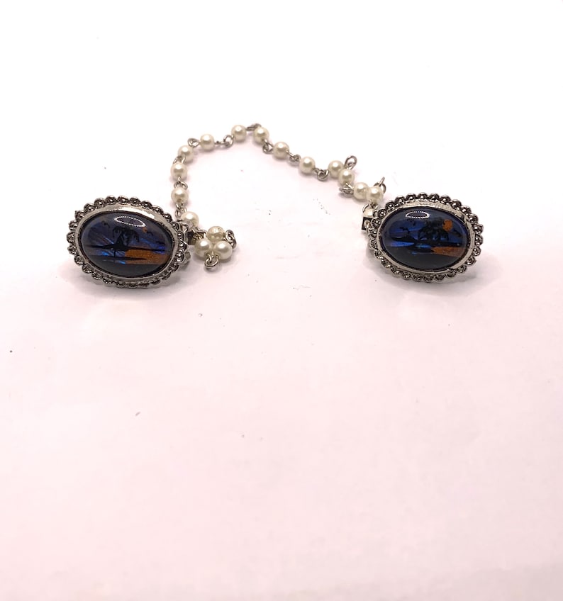 Op de afbeelding: Een paar zilverkleurige ovale broches met een blauw en oranje vlinderontwerp. De broches zijn verbonden door een ketting van nepparels.