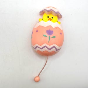 May include: An Easter-themed toy featuring a yellow chick emerging from a pink egg. The egg is decorated with purple zigzags and a purple flower. A string with a small ball hangs from the egg. The chick has a yellow beak and is peeking out of the egg.