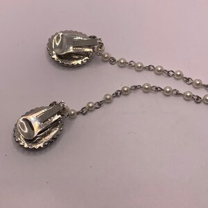 Op de afbeelding: Een paar zilveren clip-on oorbellen met een parelketting die ze verbindt. De oorbellen hebben een rond, ovaal ontwerp met een glinsterend, getextureerd oppervlak.