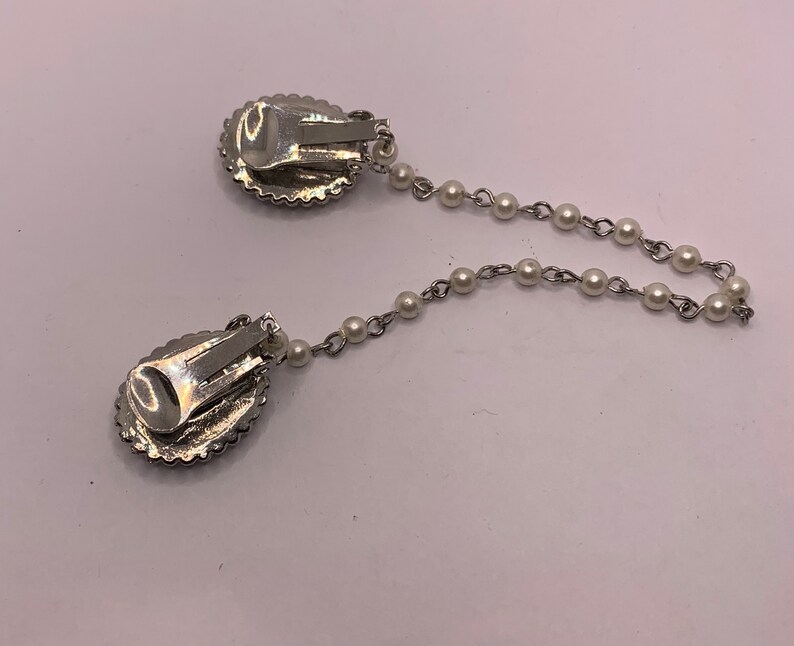Op de afbeelding: Een paar zilveren clip-on oorbellen met een parelketting die ze verbindt. De oorbellen hebben een rond, ovaal ontwerp met een glinsterend, getextureerd oppervlak.