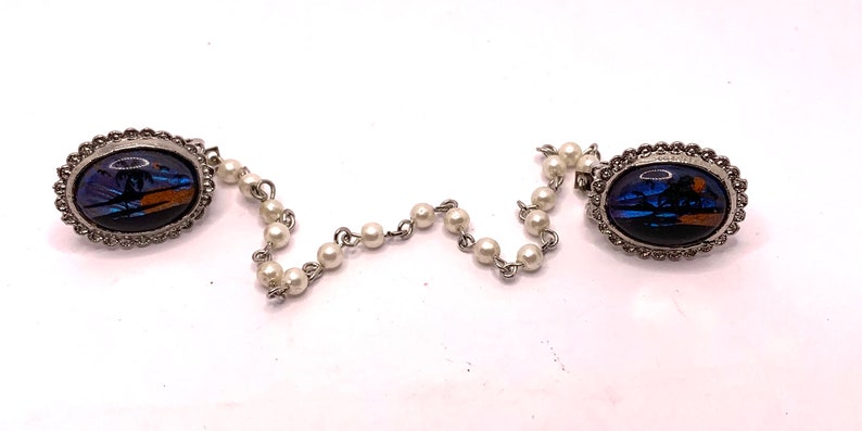Op de afbeelding: Een paar zilveren ovale broches met blauwe en oranje vlinder vleugelontwerpen verbonden door een parelketting.