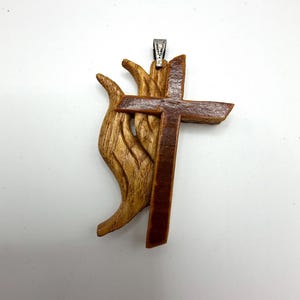 May include: Wooden pendant featuring a cross design over a stylized flame. The cross is a darker brown than the flame, and the pendant has a silver-coloured bail for hanging. The pendant is made of wood.