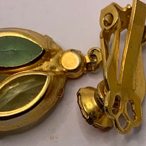 May include: A gold-tone metal clip-on earring featuring two marquise-cut stones in shades of light green and pale yellow. The earring has a hinged clip mechanism on the back and a decorative setting that holds the faceted stones securely in place.
