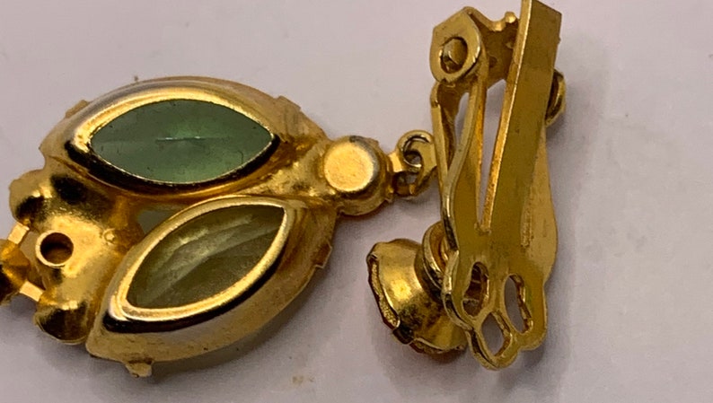 May include: A gold-tone metal clip-on earring featuring two marquise-cut stones in shades of light green and pale yellow. The earring has a hinged clip mechanism on the back and a decorative setting that holds the faceted stones securely in place.