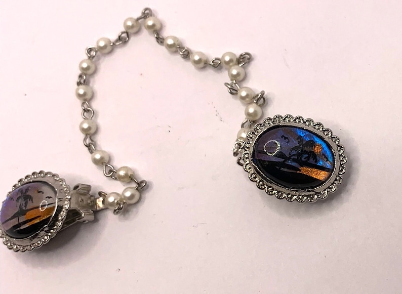 Op de afbeelding: Een paar zilverkleurige vlinder vleugel clips met een parelketting connector. De vlindervleugels zijn iriserend blauw en oranje.