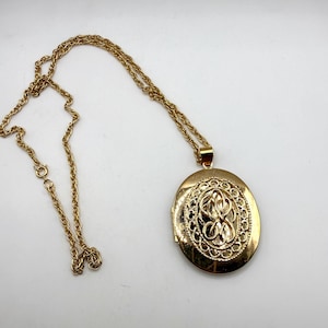 May include: Gold-toned oval locket necklace. The locket features an ornate filigree design with an intricate pattern. The chain is a delicate rope style, and the clasp is visible. The necklace is displayed on a white surface.