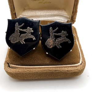 May include: A pair of vintage shield-shaped cufflinks. Each black cufflink features a silver-toned chariot scene with a figure and a horse. The cufflinks are displayed in a brown velvet lined box.