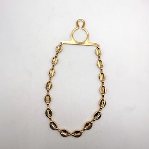 May include: A gold-toned necklace with an oval chain design. The necklace has a unique pendant with a circular top and a horizontal bar connecting to the chain. The chain is made of interconnected oval links. The jewelry is displayed against a white background.