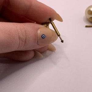 May include: Close-up of a gold earring with a pearl and a small, pointed metal post. The earring is held in a hand with a blue and white eye design on the fingernail.