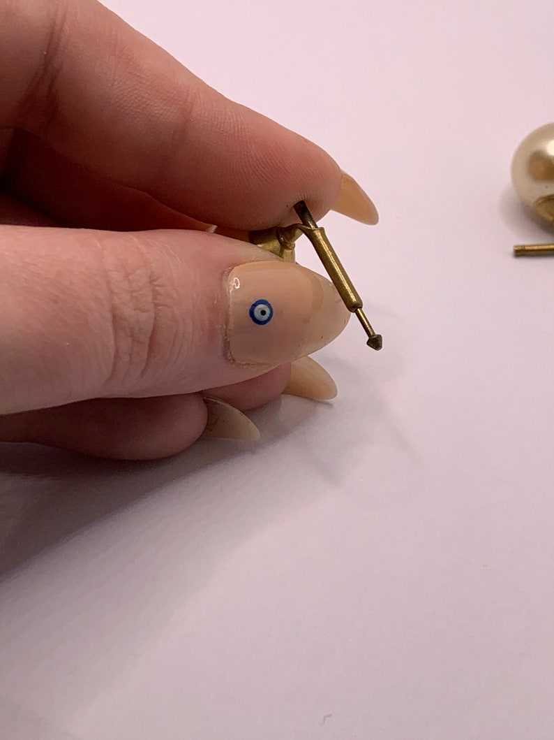 May include: Close-up of a gold earring with a pearl and a small, pointed metal post. The earring is held in a hand with a blue and white eye design on the fingernail.
