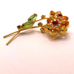 May include: A gold-tone metal brooch shaped like a flower spray with two light green marquise-cut stones as leaves. The flower heads feature clusters of orange, yellow, and clear faceted crystals with small white enamel accents at the centre.