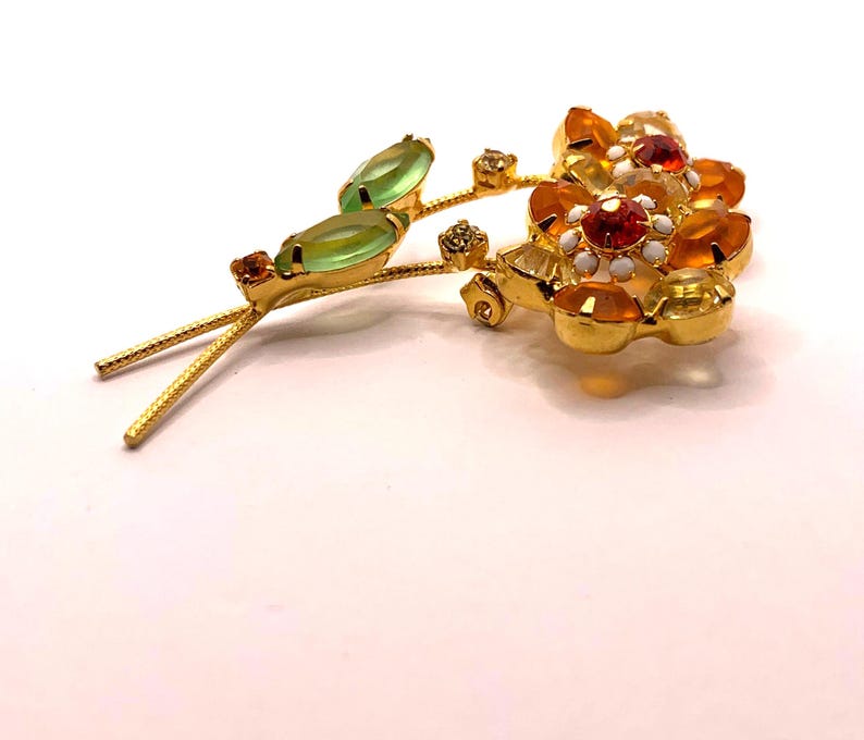 May include: A gold-tone metal brooch shaped like a flower spray with two light green marquise-cut stones as leaves. The flower heads feature clusters of orange, yellow, and clear faceted crystals with small white enamel accents at the centre.