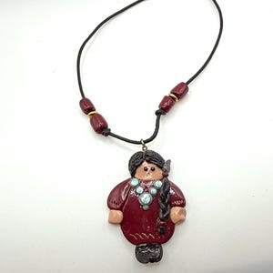 Può includere: Una collana in argilla rossa con un ciondolo che rappresenta una figura stilizzata di una donna con una lunga treccia, che indossa una collana turchese e un vestito rosso. La collana è realizzata con perline rosse e dorate su un cordoncino nero.