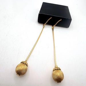 May include: A pair of long gold-tone drop earrings featuring slender stems attached to textured, oval-shaped pendants with a ribbed finish. The earrings are displayed against a small, rectangular black box.