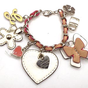Puede incluir: Un brazalete de cuero blanco con una cadena plateada y varios abalorios, incluyendo un corazón con la palabra "Guess" en él, una mariposa, una flor y un candado.