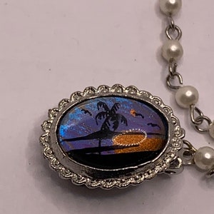 Op de afbeelding: Een zilveren ovale hangerketting met een blauw en paars vlinder vleugel ontwerp met een palmboom silhouet en een zonsondergang. De ketting is gemaakt van een parelketting.