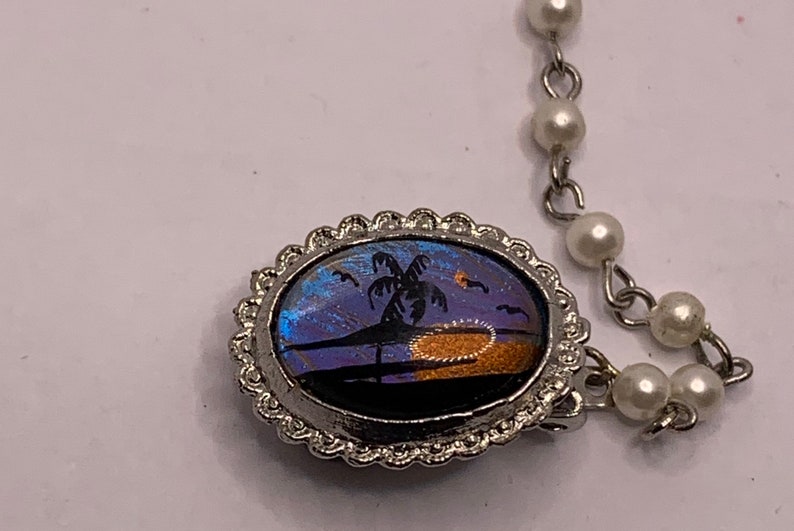 Op de afbeelding: Een zilveren ovale hangerketting met een blauw en paars vlinder vleugel ontwerp met een palmboom silhouet en een zonsondergang. De ketting is gemaakt van een parelketting.