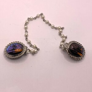 Op de afbeelding: Een paar zilverkleurige ovale broches met een parelketting die ze verbindt. De broches hebben een vlinder vleugel ontwerp met een palmboom en een zonsondergang sc&egrave;ne.