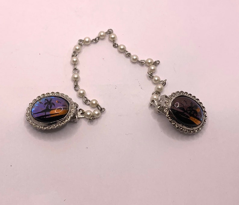 Op de afbeelding: Een paar zilverkleurige ovale broches met een parelketting die ze verbindt. De broches hebben een vlinder vleugel ontwerp met een palmboom en een zonsondergang sc&egrave;ne.