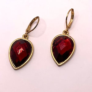 May include: A pair of gold-toned drop earrings with teardrop-shaped, faceted, deep red gemstones. The gemstones are set in a gold-colored bezel and have a lever-back closure.