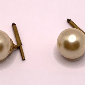 May include: A pair of pearl cufflinks with gold-toned metal posts. The pearls are round and smooth, and the posts are straight with a slight bend at the top.