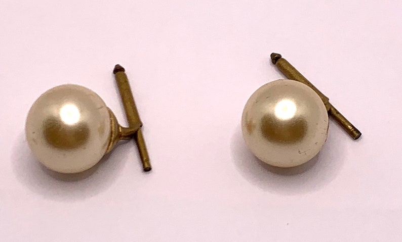 May include: A pair of pearl cufflinks with gold-toned metal posts. The pearls are round and smooth, and the posts are straight with a slight bend at the top.
