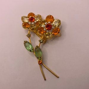 May include: A gold-tone metal brooch featuring two flower heads with orange and pale yellow faceted crystals, white enamel accents, and two green marquise-cut crystal leaves on thin stems. The design includes small orange and clear crystal details.