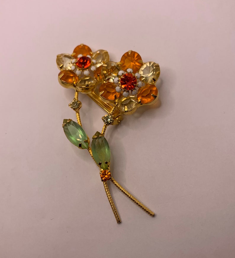 May include: A gold-tone metal brooch featuring two flower heads with orange and pale yellow faceted crystals, white enamel accents, and two green marquise-cut crystal leaves on thin stems. The design includes small orange and clear crystal details.