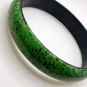 Könnte beinhalten: Ein grünes Armreif-Armband mit floralem Muster. Das Armband hat eine schwarze Innenseite und ist mit einem sich wiederholenden Muster aus grünen Blumen und Blättern auf schwarzem Hintergrund verziert. Der Armreif hat einen Durchmesser von ca. 7,6 cm.